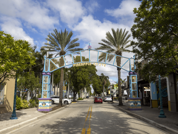 A colorful arched sign reading Pineapple Grove spans across a quiet street lined with palm trees, shops, and parked cars under a bright, partly cloudy sky.