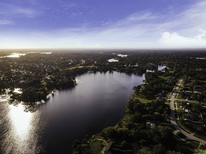 Aerial view of a suburban neighborhood beside a large lake with tree-lined streets, houses, and a vast sky with scattered clouds illuminated by sunlight.
