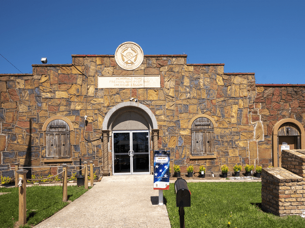 A brown stone building with two wooden-shuttered windows and a central glass door under a sign reading “American Legion.” A mailbox and flag retirement box are near the entrance, with manicured grass and clear blue sky.