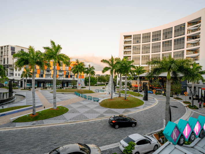A modern urban plaza with palm trees, curved roads, benches, and parked cars, surrounded by contemporary multi-story buildings during daylight.