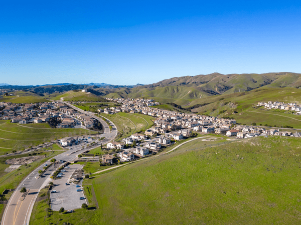 Aerial view of a suburban neighborhood with rows of houses surrounded by rolling green hills and clear blue sky. A winding road runs through the community, and a parking lot is visible in the foreground.