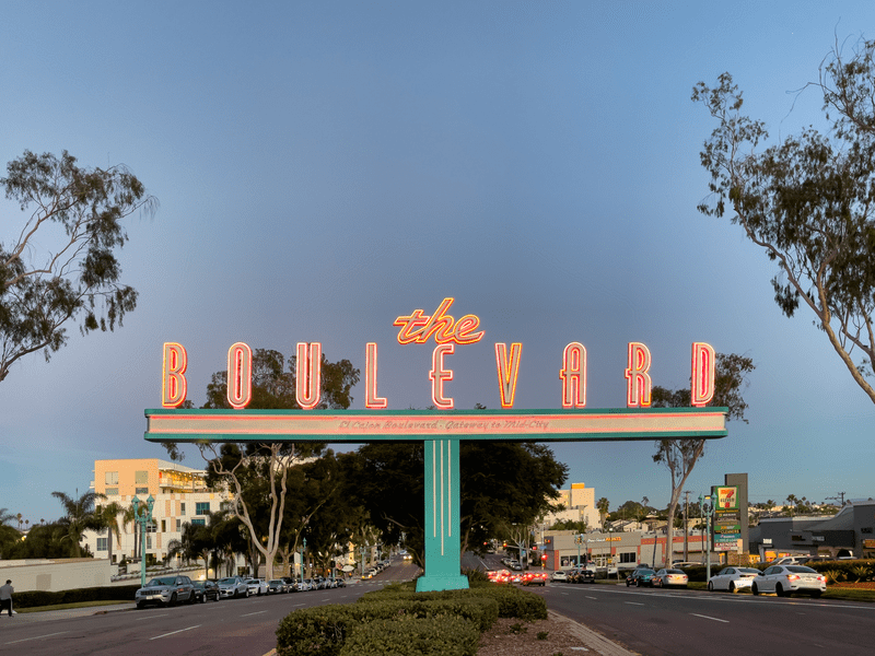 A neon sign reading The Boulevard spans across a busy street lined with cars and trees, with buildings and shops visible in the background under a clear blue sky.