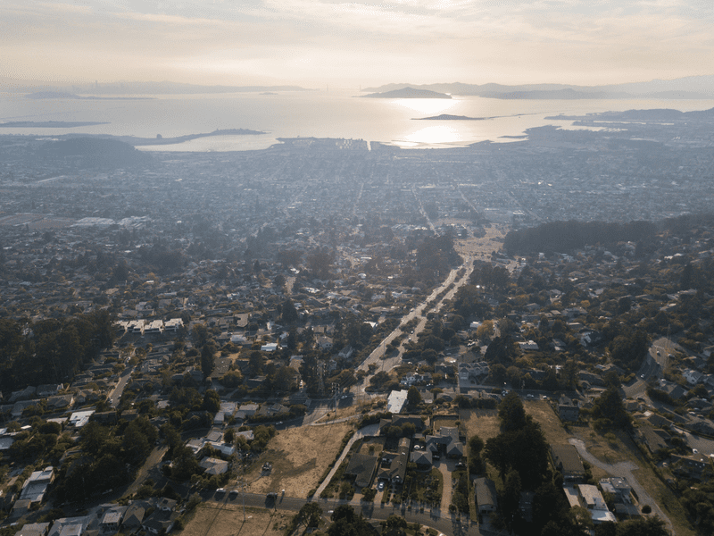 Aerial view of a suburban neighborhood with winding roads and houses, overlooking a body of water at sunset. The sun reflects off the water, with distant hills and islands visible under a hazy sky.