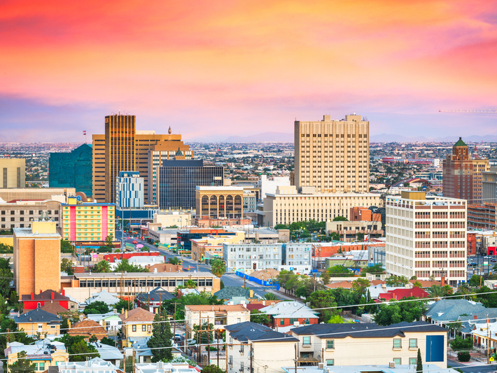 Colorful sunset sky over a city skyline with tall buildings, mid-rise structures, and residential houses in the foreground, creating a vibrant urban landscape.