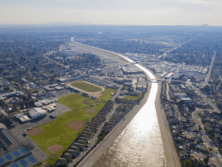 Aerial view of an urban area with a wide concrete river, baseball fields, green lawns, residential neighborhoods, and a school complex under a hazy sky.
