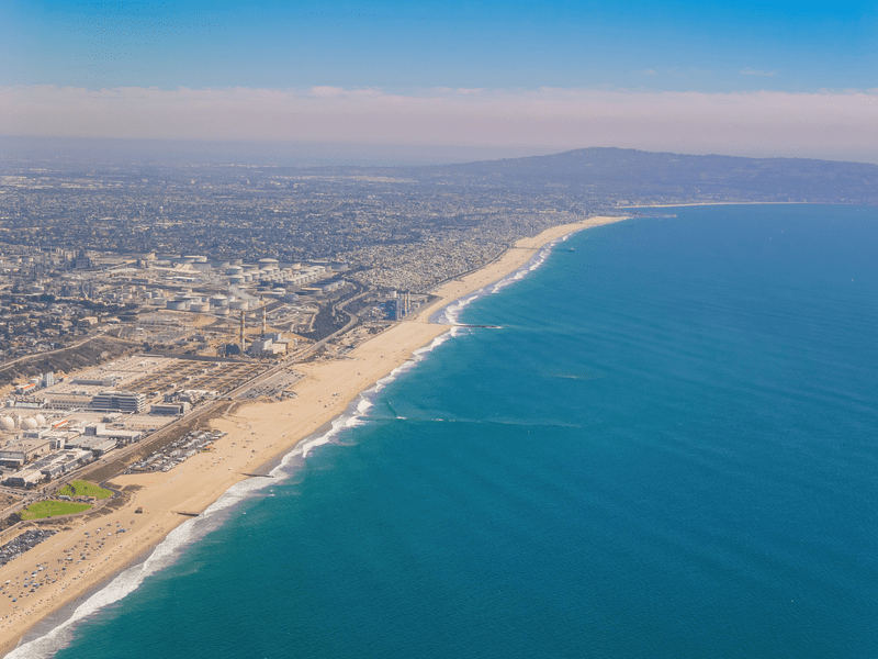 Aerial view of a long sandy beach with gentle waves, a large city in the background, and mountains in the distance under a clear blue sky.
