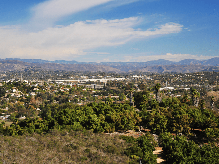 A scenic view of a suburban area with green trees in the foreground, houses scattered throughout, and mountains in the background under a partly cloudy blue sky.