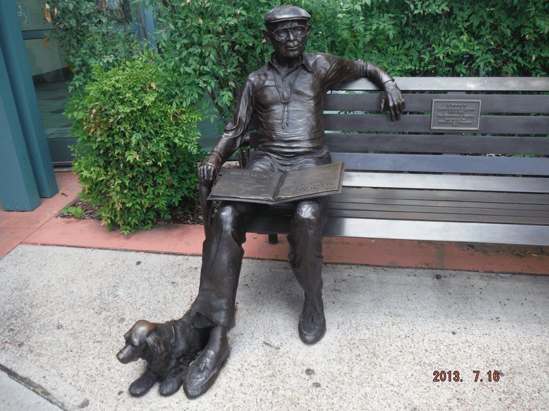 A bronze statue of a man sitting on a bench with an open book on his lap and a dog lying at his feet, surrounded by greenery; date stamp reads 2013. 7. 16.