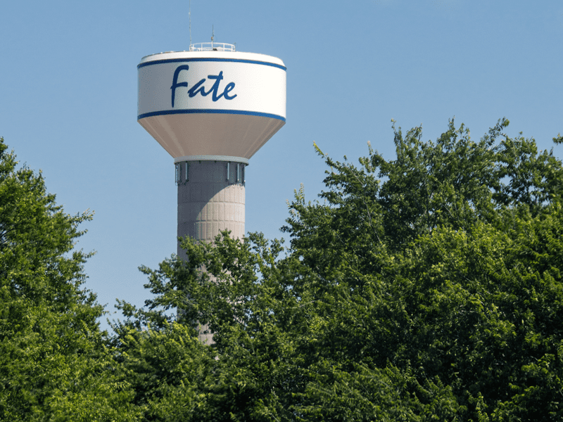 A tall water tower with the word Fate painted on the tank rises above green leafy trees against a clear blue sky.