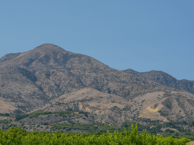 A dry, rugged mountain range with sparse vegetation rises above a green foreground of trees under a clear blue sky.