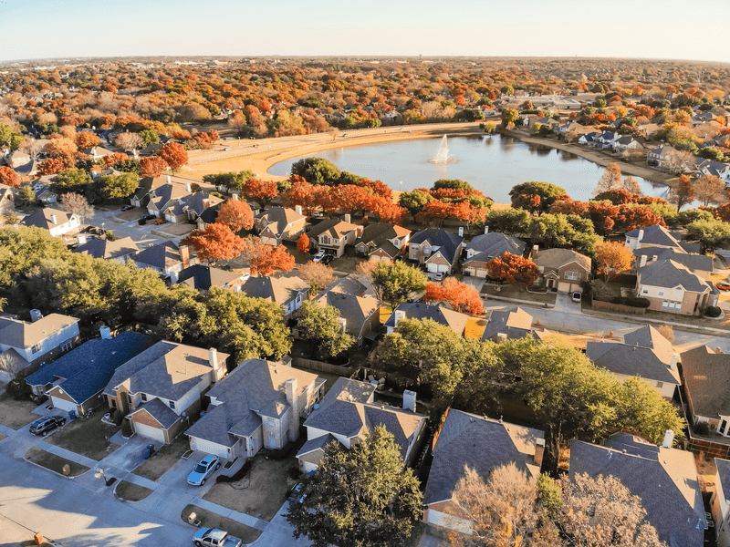 Aerial view of a suburban neighborhood with houses, tree-lined streets, and a small lake with a fountain in the center, surrounded by colorful autumn foliage.