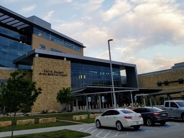 Exterior view of Carl R. Darnall Army Medical Center, a modern multi-story building with large windows. Several cars are parked in front, and trees and a blue sky with clouds are visible.