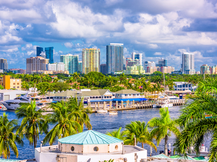 A waterfront cityscape featuring tall modern buildings, palm trees, boats docked along the water, and a vibrant, partly cloudy sky in the background.