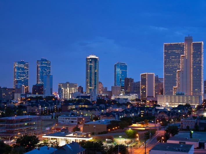 A city skyline at dusk with several tall buildings outlined in lights, set against a deep blue sky. Streets and smaller buildings are visible in the foreground, illuminated by streetlights.