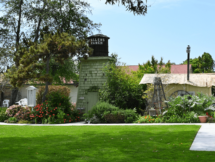 A lush garden with green grass, vibrant flowers, and tall trees surrounds a small green building with a water tower labeled ALLENS RANCH. A rustic house and garden structures are visible in the background.