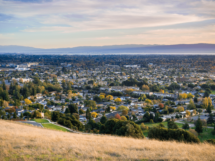 A panoramic view of a suburban city with houses, trees, and buildings, seen from a hillside with dry grass under a partly cloudy sky and distant mountains in the background.