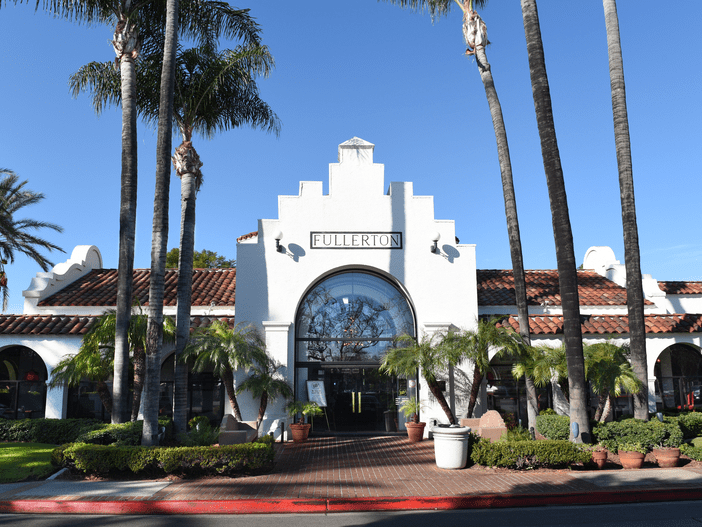 A white stucco building with a red-tiled roof, arched windows, palm trees, and a sign reading “Fullerton” above the main entrance. The sky is clear and blue.