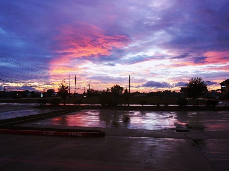 A mostly empty parking lot with wet pavement reflecting a dramatic sunrise or sunset sky filled with vibrant pink, purple, and blue clouds. Power lines and a few trees are visible in the background.