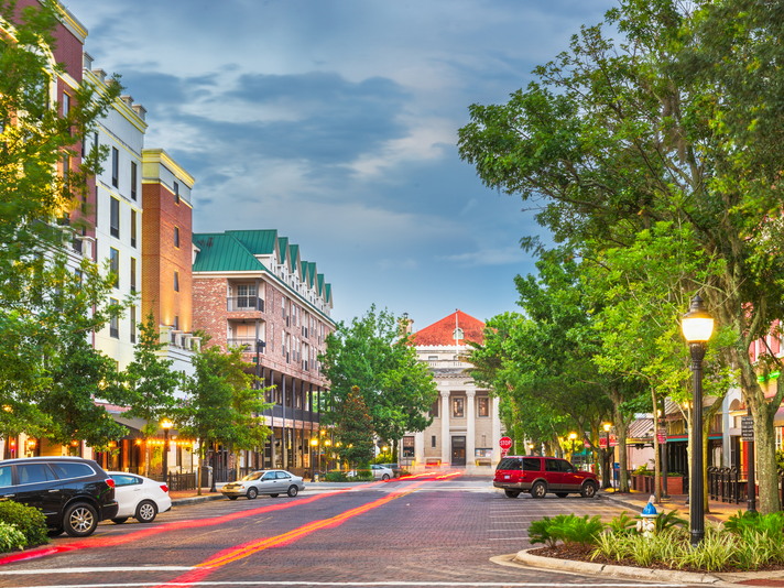 A vibrant, tree-lined street in a small city with brick buildings, parked cars, and a historic white building with columns at the end of the street under a cloudy blue sky.