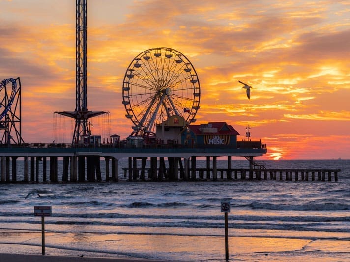 A ferris wheel and amusement park sit on a pier over the ocean at sunset, with vibrant orange and yellow skies, gentle waves, and a seagull flying near the pier.
