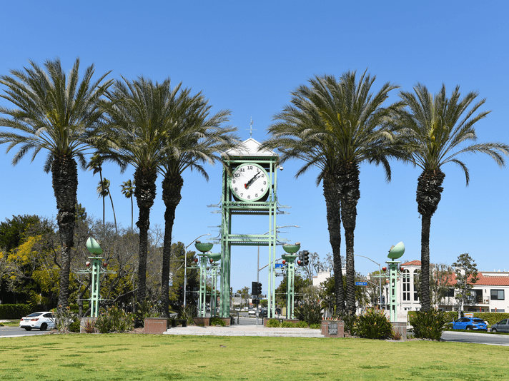 A large outdoor clock structure surrounded by tall palm trees stands on a grassy area near a road, with cars and buildings visible in the background under a clear blue sky.