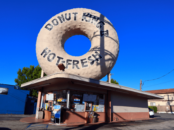 A donut shop with a giant beige donut sculpture on the roof that reads Donut King and Hot Fresh against a clear blue sky. The shop has large windows and a menu displayed outside.