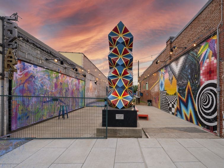 A colorful geometric sculpture stands in an outdoor art alley, surrounded by vibrant murals on brick walls and string lights overhead, with a dramatic sunset sky in the background.