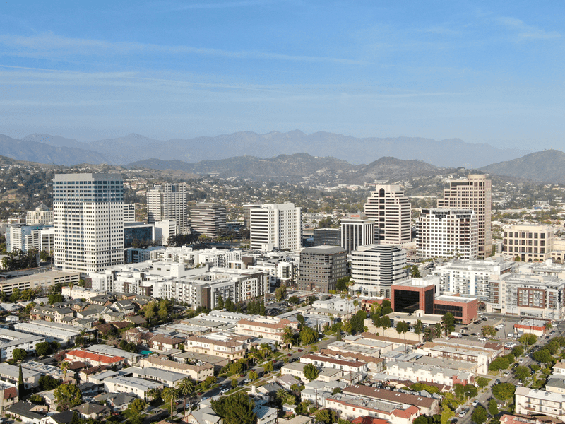 Aerial view of a cityscape with tall office buildings, residential areas, and tree-lined streets, set against a backdrop of mountains under a clear blue sky.