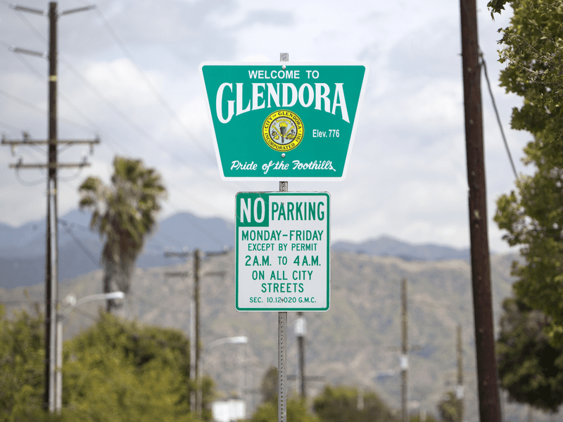 A green sign welcomes visitors to Glendora, Pride of the Foothills, above a white sign stating no parking Monday to Friday, 2 a.m. to 4 a.m., except by permit, with mountains and trees in the background.