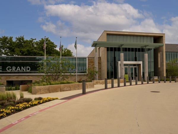 Modern building with large glass windows, stone walls, and a covered entrance. Three flagpoles stand nearby, and landscaping with flowers and bushes borders a curved driveway in front.
