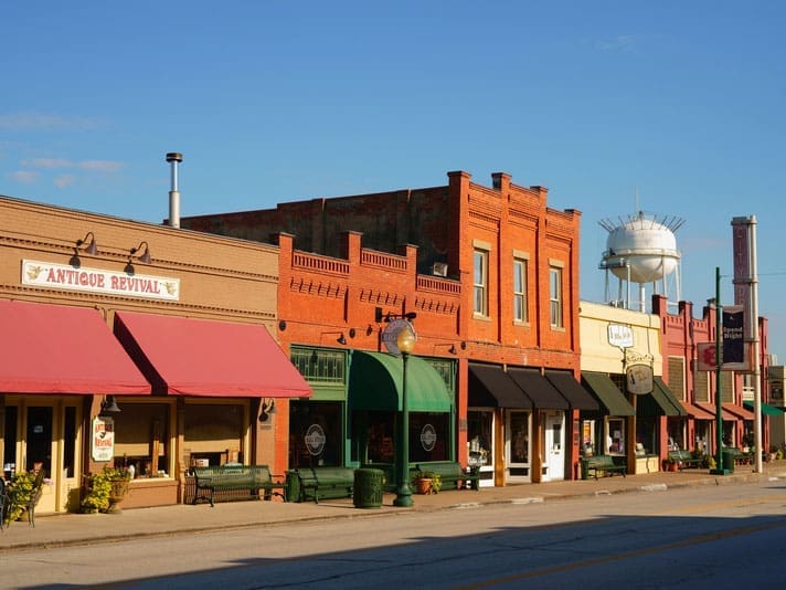 A small-town main street with historic brick buildings, colorful awnings, antique and café signs, benches, and a water tower in the background under a clear blue sky.