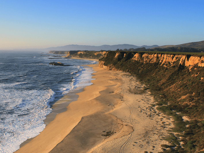 Aerial view of a sandy beach bordered by ocean waves on one side and steep cliffs and greenery on the other, under a clear blue sky at sunset.