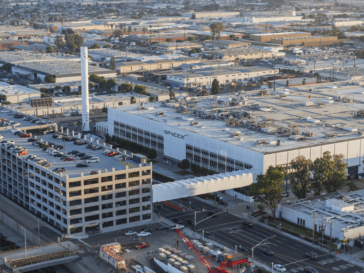 Aerial view of the SpaceX headquarters in Hawthorne, California, showing the large white building with the SpaceX logo, adjacent parking structure, and surrounding industrial area.