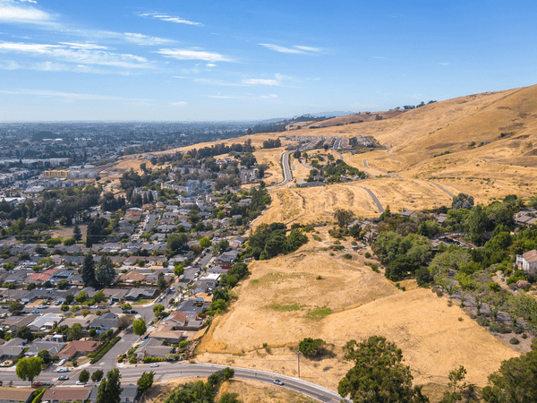Aerial view of a suburban neighborhood bordering golden, grassy hills under a blue sky, with houses clustered on one side and open, rolling landscape on the other.
