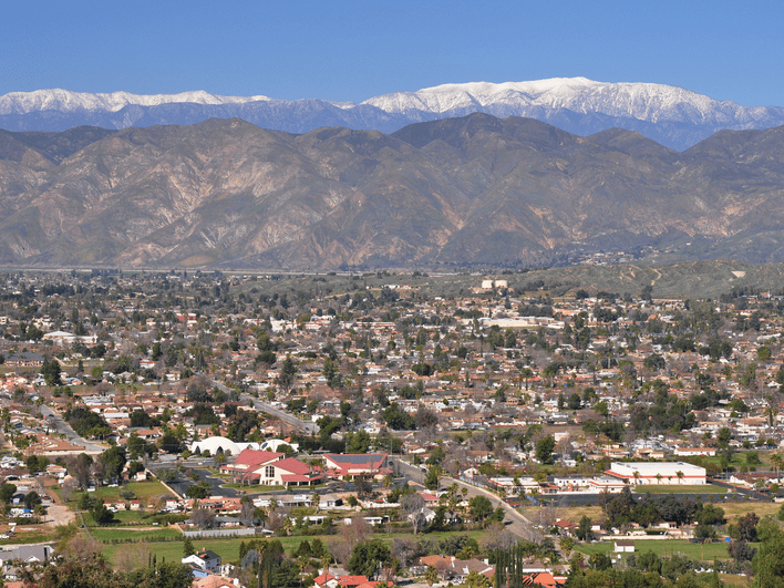 A wide view of a suburban city with many houses and buildings, set against a backdrop of tall, rugged mountains topped with snow under a clear blue sky.