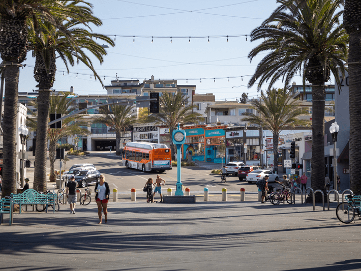 A sunny street scene with palm trees, a turquoise clock, parked cars, a city bus, colorful shops, and people walking or biking in a relaxed, coastal town atmosphere.