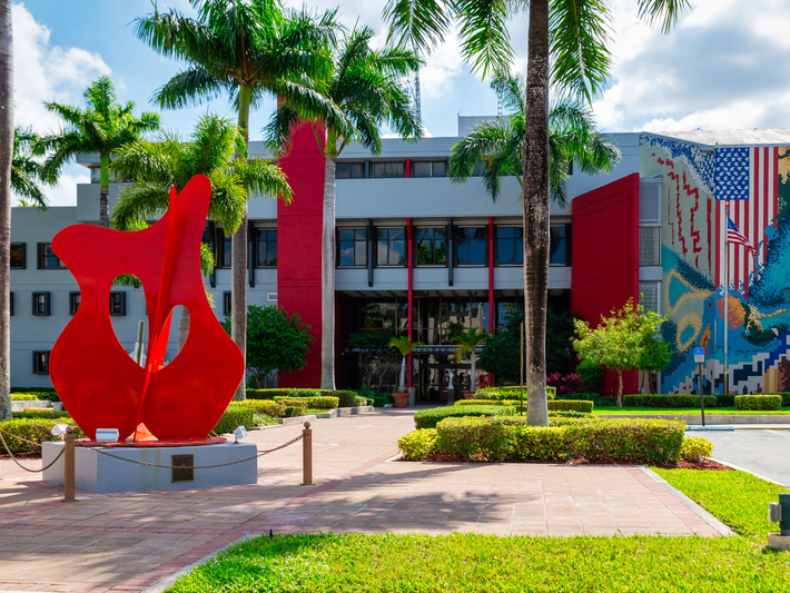 A bright red abstract sculpture stands in front of a modern building with large windows, red columns, palm trees, neatly trimmed bushes, and a colorful mural featuring the American flag on one wall.