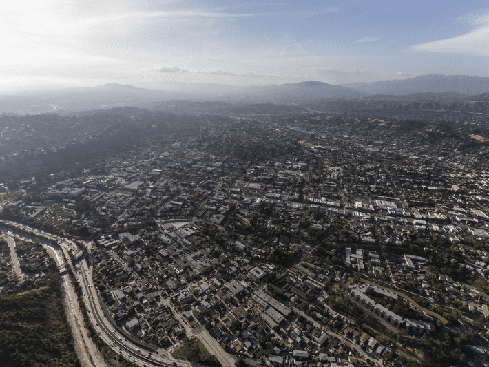 Aerial view of a sprawling cityscape with widespread buildings, roads, and highways, surrounded by distant mountains under a hazy sky.