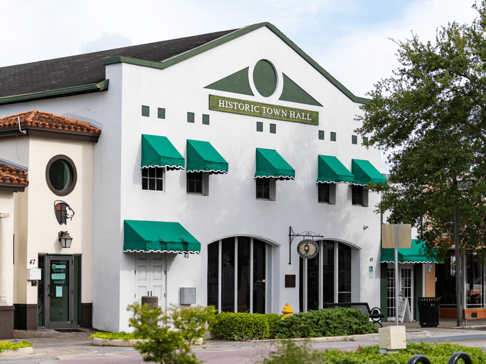 A white, historic town hall building with green awnings over windows and doors, arched upper windows, and a sign reading Historic Town Hall above the entrance. Trees and neighboring buildings are visible nearby.