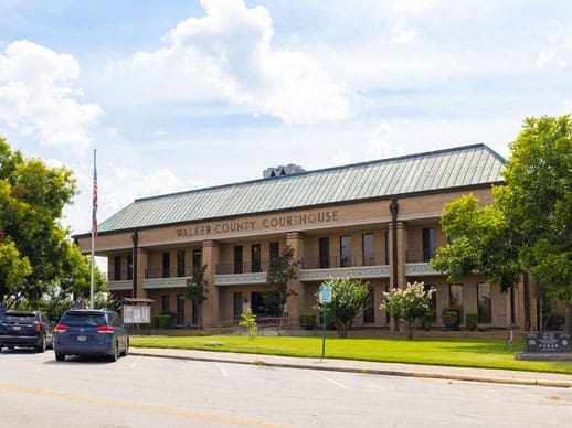 A two-story brick building with a green roof labeled Walker County Courthouse, surrounded by trees and cars parked along the street on a sunny day.