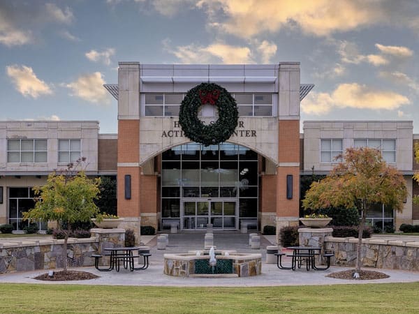 A modern activity center with large glass doors, a wreath above the entrance, and a small fountain surrounded by picnic tables in front. Trees with autumn foliage line the walkway. The sky is partly cloudy.