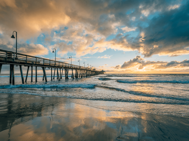 A long wooden pier stretches over the ocean at sunset, with dramatic clouds and golden sunlight reflected in the wet sand and gentle waves below.