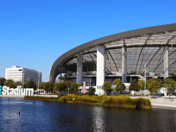 A large modern stadium with an open roof is seen beside a pond, with green trees and a clear blue sky in the background. The word Stadium appears on a large sign near the water.