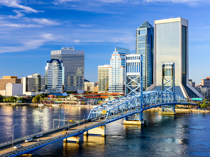 A blue steel bridge crosses a river in front of a modern city skyline with tall glass buildings under a bright, partly cloudy sky.
