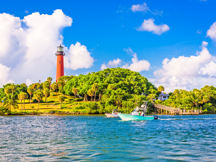 A red lighthouse stands on a green, tree-covered hill near the shoreline, with a white boat cruising on blue water under a bright sky with scattered clouds.