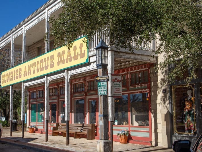 A historic two-story building with a large yellow “Sunrise Antique Mall” sign, red-trimmed windows, benches out front, and a streetlamp with parking signs in a sunny, tree-lined area.