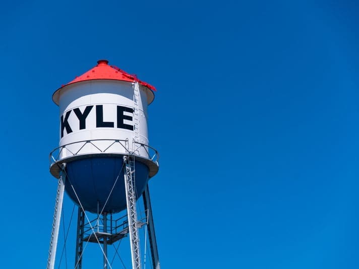 A tall white water tower with a red roof and the word KYLE written in large black letters stands against a clear blue sky.