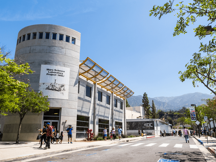 People walk near a modern, cylindrical building labeled Mars Science Laboratory Project (MSL) on a sunny day, with mountains and trees visible in the background.