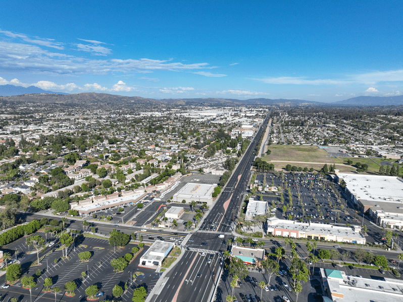 A wide aerial view of a cityscape shows a long, busy road stretching into the distance, lined with trees, buildings, parking lots, and residential areas under a bright blue sky with scattered clouds.