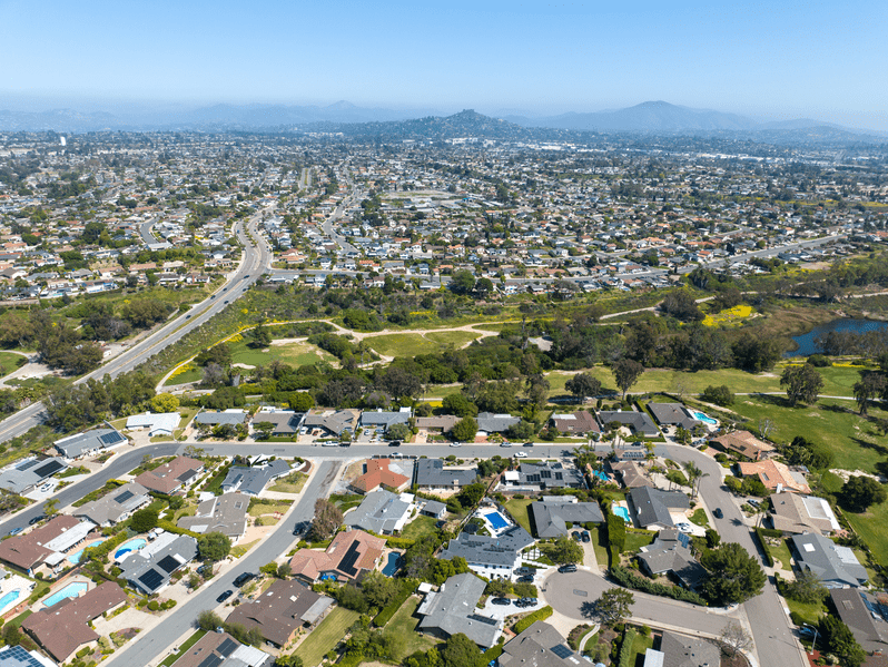 Aerial view of a suburban neighborhood with rows of houses, tree-lined streets, a main road, and distant hills under a clear blue sky. The landscape is green with scattered patches of open space.